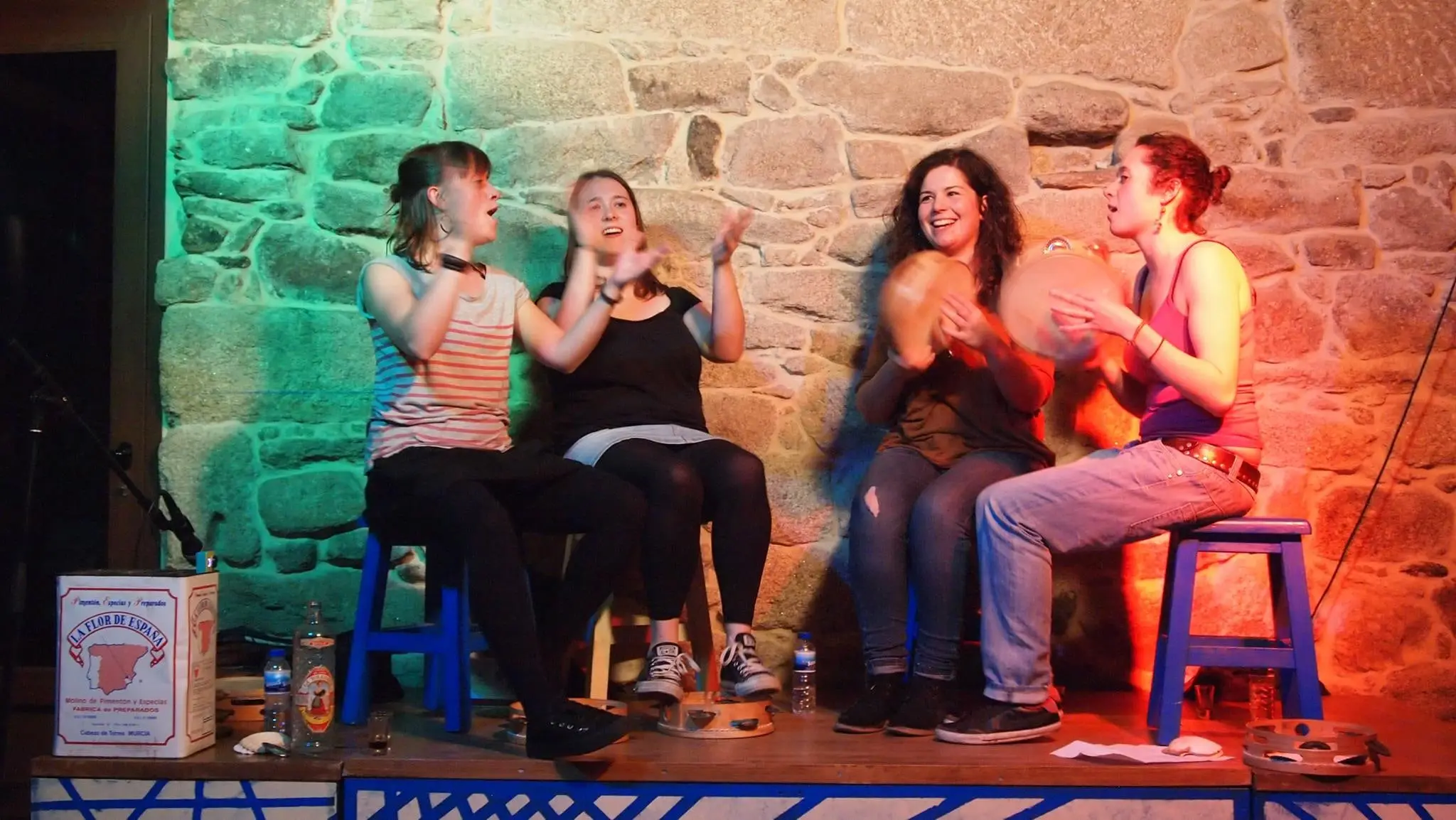 Catro mulleres cantan e baten palmas sentadas nun escenario, con luces verdes e laranxas