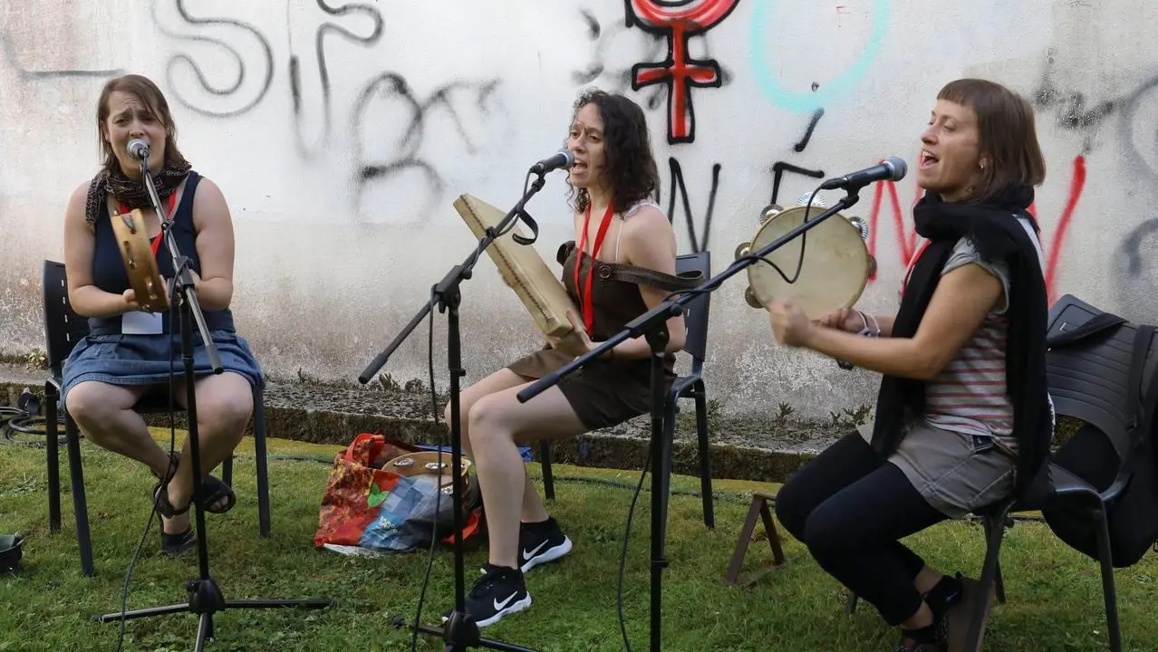 Tres pandeireteiras tocan sentadas na herba, diante dun muro con pintadas